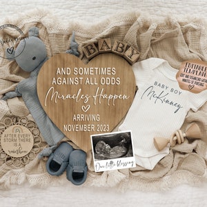 May include: A wooden heart-shaped sign with the text "And sometimes against all odds miracles happen" and "Arriving November 2023". A white onesie with the text "Baby Boy McKinney" and a black and white ultrasound image are also included in the image.