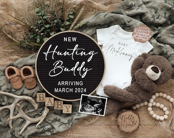 May include: A black and white letter board with the text "New Hunting Buddy Arriving March 2024". A white onesie with the text "Baby McKinney" is next to the letter board. A brown teddy bear, wooden blocks spelling "Baby", a wooden sign with the text "Hello World", and a brown antler are also in the image.