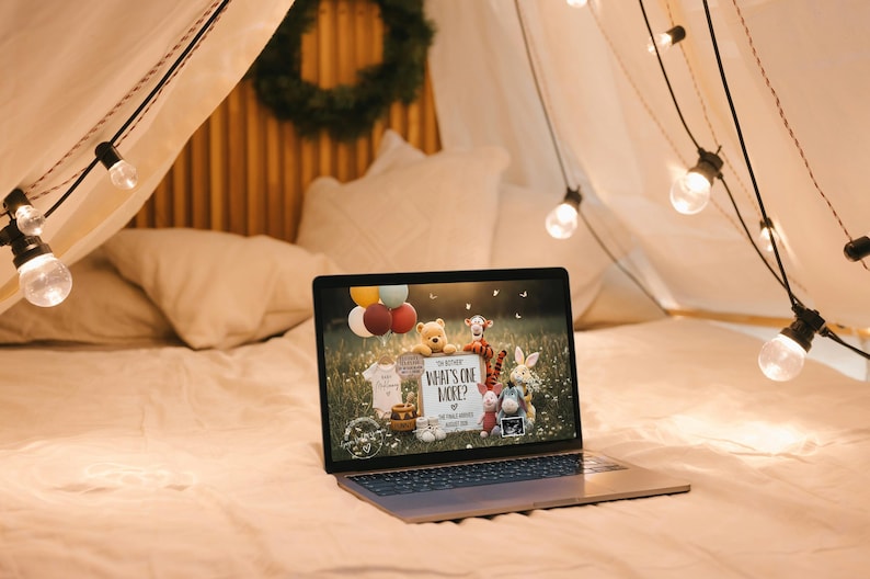 May include: A laptop displaying a baby shower invitation with Winnie the Pooh characters, balloons, and the text "What's One More?". The laptop is on a bed, surrounded by fairy lights and pillows.