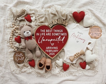 Könnte beinhalten: Ein rotes herzförmiges Schild mit dem Text "The best things in life are sometimes unexpected" und "Arriving February 2025". Neben dem Schild liegt ein weißer Strampler mit dem Text "Baby McKinney". Es gibt mehrere rote und weiße Herzen, einen gehäkelten Teddybär und ein Holzrassel.