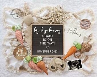 May include: A baby announcement with a letter board that reads "hip hop hooray a baby is on the way! November 2023". The announcement also includes a white onesie with the text "Baby McKinney", a wooden sign that says "Oh Baby!" and a few carrot-shaped toys.