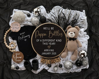 May include: A black and white baby announcement with a sign that reads "We'll be Poppin' Bottles of a Different Kind This Year! Arriving January 2025". The sign is surrounded by a white blanket with black stars, a crocheted teddy bear, wooden blocks that spell out "2025", and silver and black ornaments.