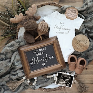 May include: A baby announcement with a white onesie that says "Baby McKinney November 2023", a wooden sign that says "Coming Soon", a letter board that says "Our Next Great Adventure", a stuffed moose, a pair of brown moccasins, and a sonogram photo.
