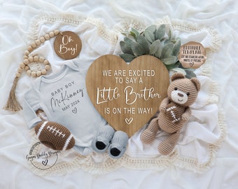 May include: A baby announcement featuring a light blue onesie with "Baby Boy McKinney May 2024" printed on it, a wooden heart sign that says "We are excited to say a Little Brother is on the way!", and a crochet football and teddy bear.