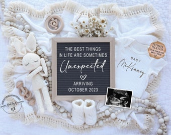 May include: A white letter board with the text "The best things in life are sometimes unexpected" and a heart symbol. Below the text is "Arriving October 2023". The letter board is surrounded by white baby booties, a white crocheted bunny, a wooden rattle, and a white onesie with the text "Baby McKinney".