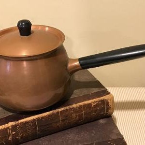 May include: A copper pot with a black handle and lid. The pot is sitting on a stack of old books.