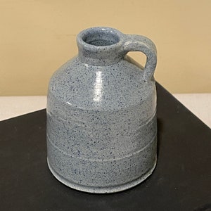 May include: A small, light blue ceramic pitcher with a speckled glaze and a single handle. The pitcher has a rounded body and a slightly flared lip.