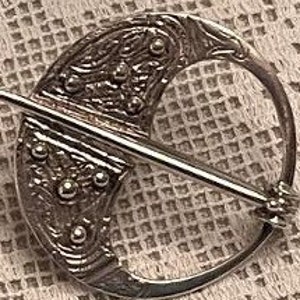 May include: A silver-toned brooch with a crescent moon design and a straight pin. The brooch features intricate embossed patterns and small, raised circular details. The pin is straight and passes through the circular frame.