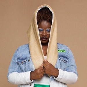 May include: A person wearing a beige hooded scarf with a white lining. The scarf is pulled up over their head, and they are wearing a blue denim jacket with a green NASA patch on the left side.