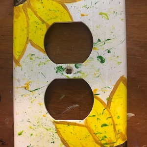 Puede incluir: Una tapa de tomacorriente blanca con un diseño pintado de dos girasoles amarillos con centros marrones. El fondo es blanco con salpicaduras de pintura verde y amarilla.