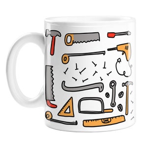 May include: White ceramic mug featuring a colourful illustration of various tools, including a hammer, saw, screwdriver, wrench, nails, and a ruler. The tools are drawn in a simple, cartoon style.