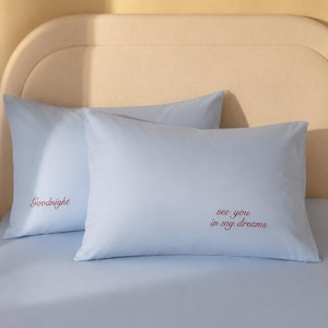 May include: Two pale blue pillowcases with red embroidered text. One says "Goodnight" and the other says "see you in my dreams". The pillowcases are on a pale blue bedsheet.