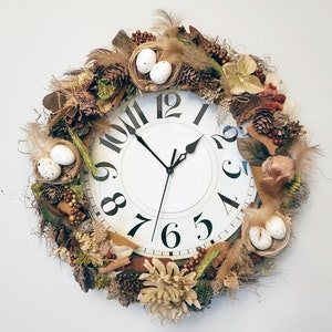 May include: A white clock with black Roman numeral markings and black hands is surrounded by a wreath of dried flowers, leaves, and twigs. The wreath includes small white eggs in nests.