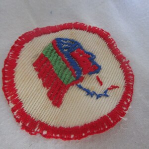 May include: A white fabric patch with a red embroidered border. The center of the patch features a red, blue, and green embroidered design that resembles a stylized head.
