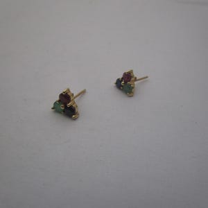 May include: A pair of gold stud earrings, each featuring a cluster of three small, round-cut gemstones in red, green, and dark blue. The stones are arranged in a triangular formation, held by gold prongs, with a thin gold post extending from the back.