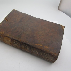 May include: A vintage, rectangular book with a worn, brown leather cover. The book's spine features embossed gold detailing. The edges of the cover show signs of wear and age, suggesting it is an antique or a well-loved item.