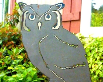 May include: A metal owl sculpture with a gray finish, perched on a wooden fence post. The owl has a detailed design with white eyes and a beak.