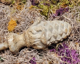 May include: A weathered, gray, decorative finial with a textured, pine cone design. The finial is lying on a bed of dried moss and other natural materials.