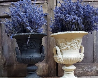 May include: Two decorative urns, one black and one cream-colored, each filled with vibrant purple lavender. The urns have ornate relief designs and handles. They sit on square bases, creating a classic, elegant display.