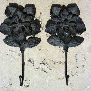 May include: Two black metal wall hooks with a floral design. Each hook features a flower with multiple petals and a leaf base, with a curved hook extending downwards. The hooks are likely for hanging items and add a decorative touch.