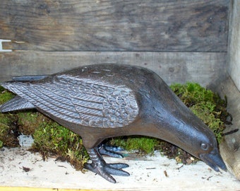 May include: A dark brown, cast iron bird figurine depicted in a foraging posture with its head lowered toward the ground. The sculpture features detailed, textured wing feathers and defined talons, showcasing a solid, heavy-duty metal construction.