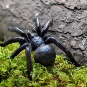May include: A black spider figurine with eight legs, a rounded abdomen, and a textured surface. The spider is positioned on a bed of green moss, with a piece of wood in the background.