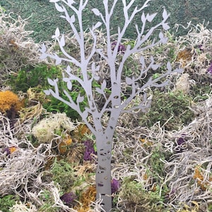 May include: A silver metal tree cutout with detailed branches and leaves, set against a backdrop of various green, yellow, and purple mosses. The tree's design is intricate, creating a natural, decorative element.