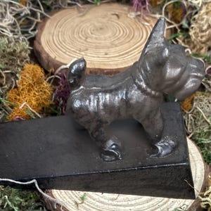 May include: A dark gray cast iron dog figurine, possibly a bulldog, standing on a rectangular base. The dog has detailed features and a textured surface. The figurine is a decorative item, likely a doorstop or bookend.