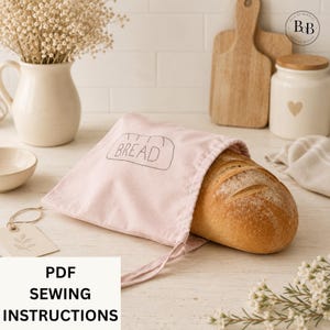 May include: A light pink fabric bread bag with a drawstring closure and the word BREAD printed in black ink. The bag contains a golden-brown loaf of crusty bread. The item is presented as a digital file for PDF sewing instructions.