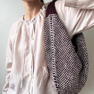Puede incluir: Bolso de hombro de punto con un patrón geométrico en blanco y burdeos. El bolso tiene forma redondeada y una correa gruesa. El fondo es una blusa rosa claro.