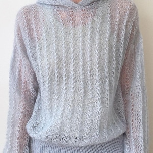 May include: A light blue, sheer, long-sleeved hooded jumper featuring a vertical open-knit pattern with a fuzzy texture. The garment has a ribbed hem and ribbed cuffs, with a hood attached at the neckline, showcasing a delicate, airy construction.