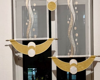 May include: Two sheer white curtains with gold and silver geometric designs. The curtains are hung on a window with a white frame. The curtains are pulled up to reveal a view of a window with a black frame. Two potted plants are on the windowsill.