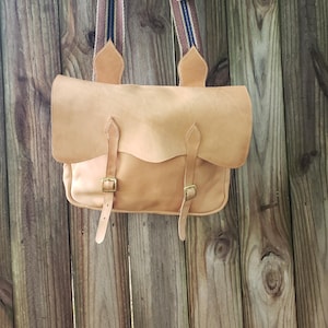 May include: A light brown leather satchel bag with a flap closure and two buckle straps. The bag has a striped shoulder strap with brown, blue, and white accents. The bag is hanging against a wooden fence.