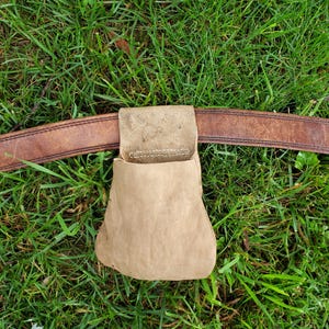 May include: A tan leather pouch attached to a brown leather belt, set against a backdrop of green grass. The pouch is rectangular with a rounded bottom. The belt is worn and shows signs of use.
