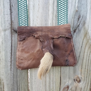 May include: A brown leather pouch with a decorative tail and a green and white strap. The pouch is made of textured leather and has white stitching along the edges. The strap is woven with a repeating pattern.