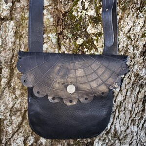 May include: A black leather bag with a spiderweb design and a silver metal accent. The bag has a scalloped edge and a long strap.