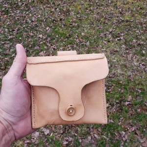 May include: A light tan leather pouch with a flap closure and a button. The pouch is held in a hand against a background of grass and fallen leaves. The pouch has a stitched edge.