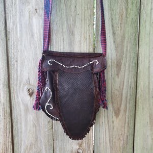 May include: A dark brown leather pouch with a textured flap and fringe detailing, adorned with white beadwork in a swirling pattern. The bag features a long, woven shoulder strap in a red and blue geometric pattern with fringed ends.