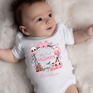May include: A white baby onesie with a pink and purple Halloween themed design. The onesie says "My First HALLOWEEN" and features a skull, a ghost, a pumpkin, and a haunted house. The name "Amelia" is written below the design.