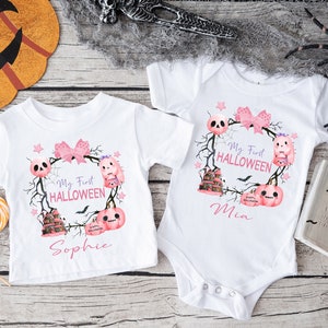May include: Two white baby onesies with pink and purple Halloween themed designs. One onesie says "My First Halloween" with a pink bow, a skull, a pumpkin, and a ghost. The other onesie says "My First Halloween" with a pink bow, a skull, a pumpkin, and a ghost. The onesie on the left says "Sophie" and the onesie on the right says "Mia".