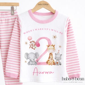 May include: A white long-sleeve shirt with pink striped sleeves and a pink collar, featuring a large pink number 2, a monkey, an elephant, a giraffe, and a zebra. The text reads WHEN I WAKE UP I WILL BE and the name Aurora in pink script.