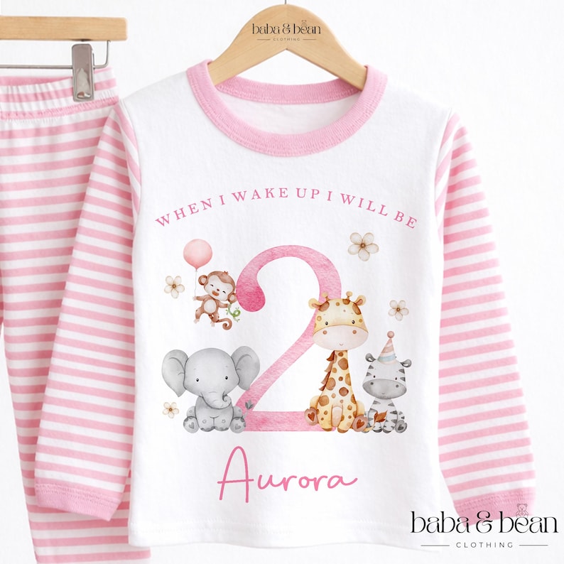 May include: A white long-sleeve shirt with pink striped sleeves and a pink collar, featuring a large pink number 2, a monkey, an elephant, a giraffe, and a zebra. The text reads WHEN I WAKE UP I WILL BE and the name Aurora in pink script.