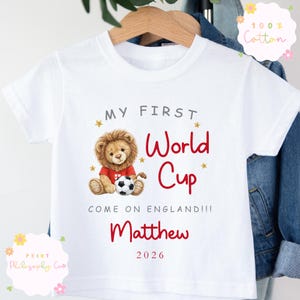 May include: White t-shirt with the text "MY FIRST World Cup" in grey and red. A cartoon lion wearing a red shirt and holding a football is featured. The name "Matthew" and the year "2026" are also printed on the shirt.