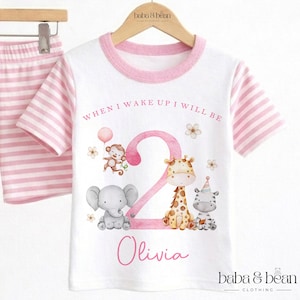 May include: A white t-shirt with pink striped sleeves and a pink collar, featuring a large pink number 2, illustrations of a monkey, elephant, giraffe, and zebra, and the text "WHEN I WAKE UP I WILL BE" and the name "Olivia" in pink.
