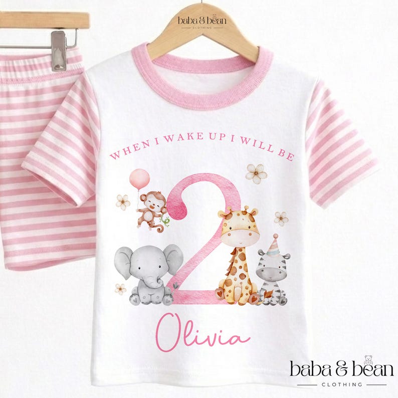 May include: A white t-shirt with pink striped sleeves and a pink collar, featuring a large pink number 2, illustrations of a monkey, elephant, giraffe, and zebra, and the text "WHEN I WAKE UP I WILL BE" and the name "Olivia" in pink.