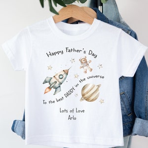 Op de afbeelding: Wit T-shirt met een raket, een planeet, een teddybeer-astronaut en sterren. De tekst luidt "Happy Father's Day To the best Daddy in the universe Lots of Love Arlo".