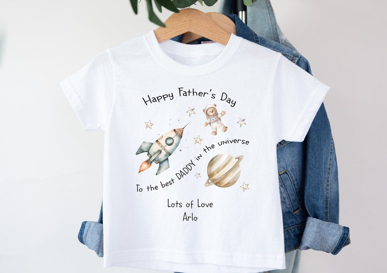 Op de afbeelding: Wit T-shirt met een raket, een planeet, een teddybeer-astronaut en sterren. De tekst luidt "Happy Father's Day To the best Daddy in the universe Lots of Love Arlo".