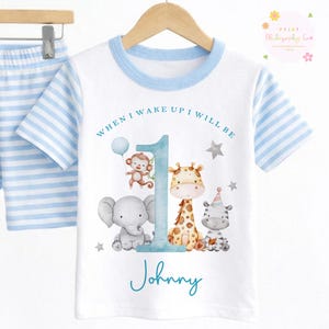 May include: A white and blue striped short-sleeve pajama set. The white t-shirt has a blue collar and features a number 1 with a monkey, elephant, giraffe, and zebra. The text reads "When I wake up I will be" and "Johnny".