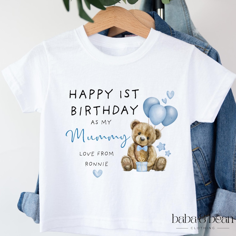 Pode incluir: Uma t-shirt branca de manga curta com uma ilustra&ccedil;&atilde;o de um urso de peluche castanho, bal&otilde;es azuis e o texto: HAPPY 1ST BIRTHDAY AS MY Mummy LOVE FROM RONNIE. O design inclui pequenos cora&ccedil;&otilde;es e estrelas azuis &agrave; volta do urso.