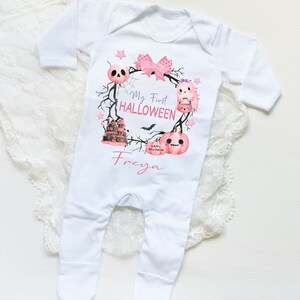 May include: White baby onesie with a pink and purple Halloween design. The design features a wreath with a skull, a ghost, a house, and pumpkins. The text "My First Halloween" and the name "Freya" are printed on the onesie.